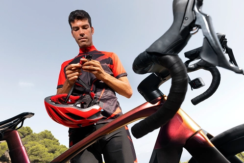 A male cyclist in a red and black jersey fastening his red helmet while standing next to his road bike.