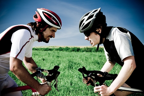 Two male cyclists in helmets looking at each other and shouting aggressively or competitively while riding on a green field background.